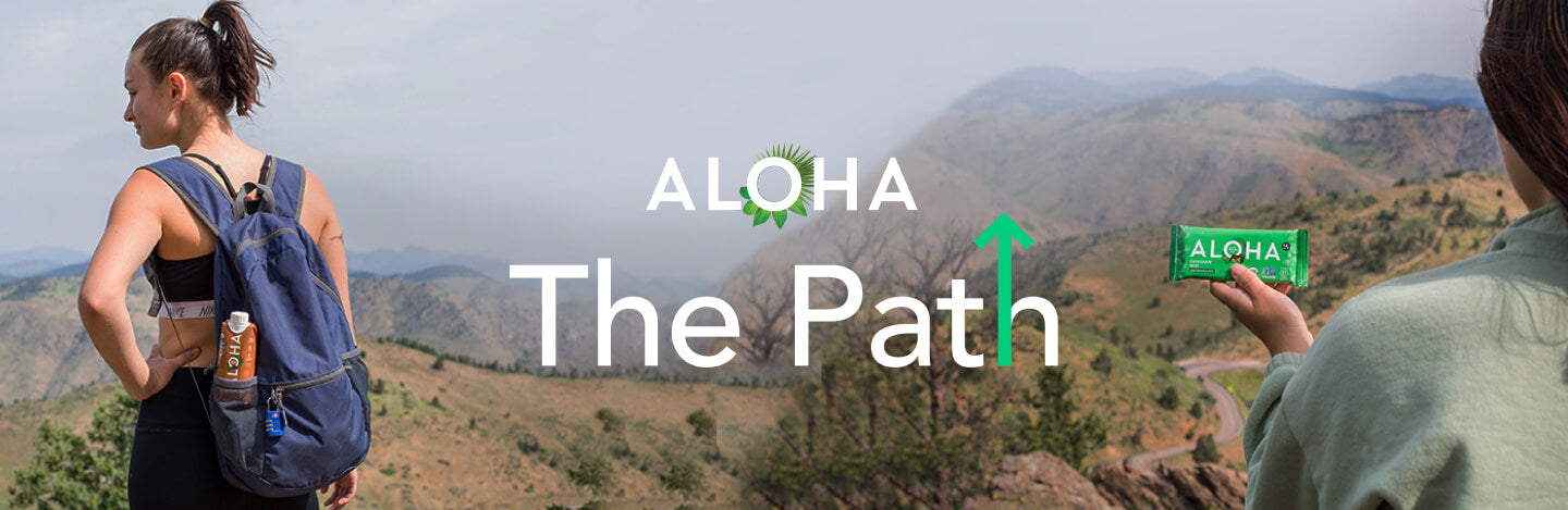 Aloha The Path: Woman standing in front of mountain view with Aloha protein shake in her backpack. Another person is holding an Aloha protein bar.