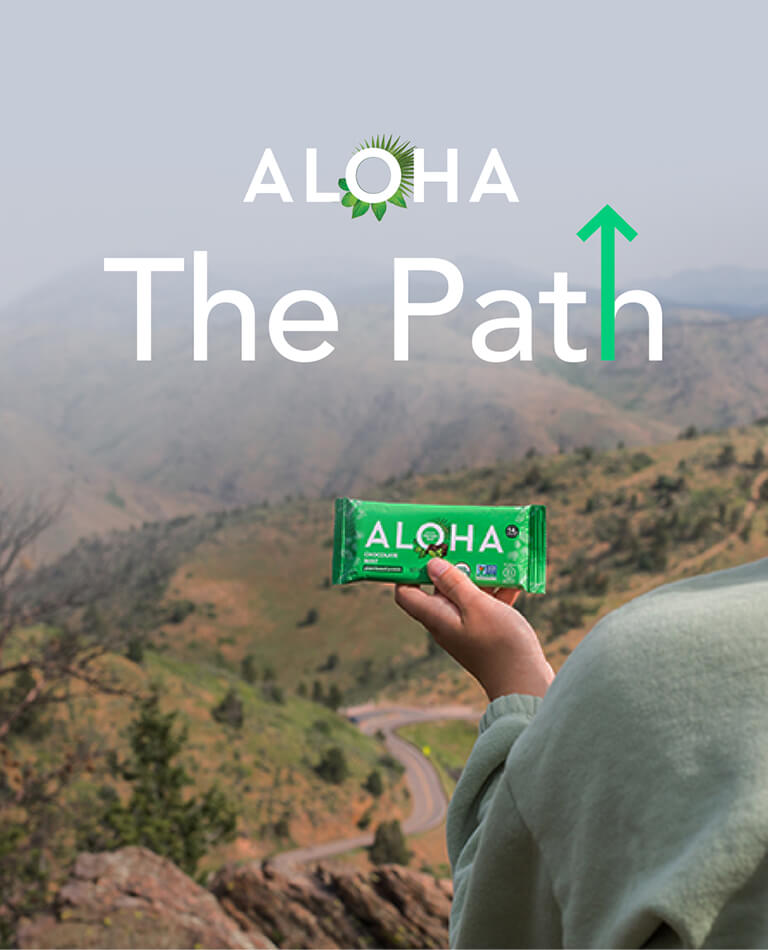 Aloha The Path: Person in front of mountain view holding an Aloha protein bar.
